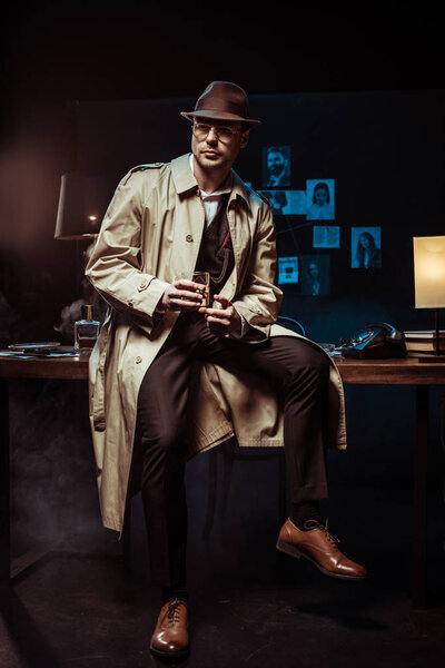 Detective in hat and trench coat sitting on table with glass of cognac