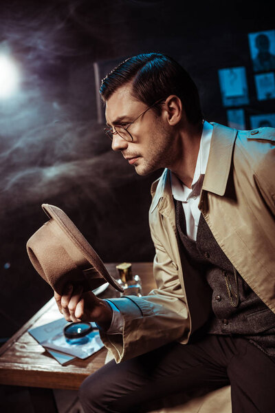 Pensive detective in glasses and trench coat sitting on table and holding hat