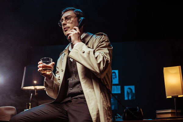 Low angle view of detective in glasses talking on telephone and holding glass of cognac