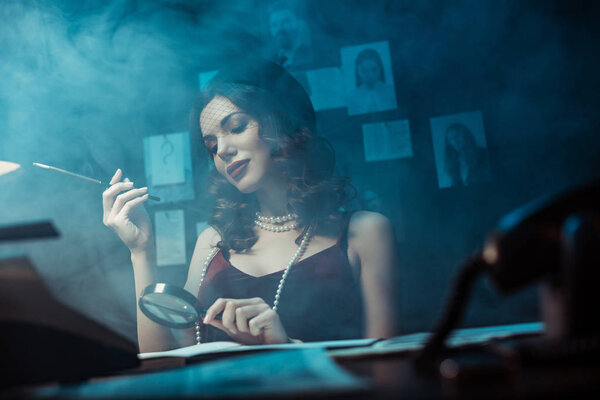 Привлекательная женщина в шляпе-фасцинаторе держит мундштук и увеличитель в темном кабинете
