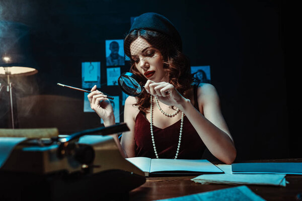 Привлекательная женщина в шляпе-фасцинаторе держит мундштук и увеличитель в темном кабинете
