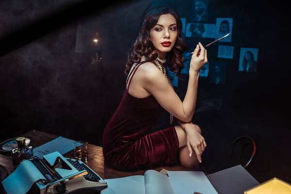 Красивая молодая женщина в платье держит мундштук, сидя на столе в темном офисе

