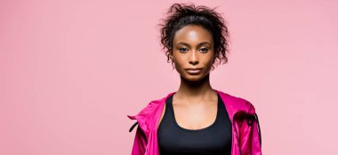 panoramic shot of attractive african american sportswoman in windbreaker looking at camera isolated on pink with copy space clipart