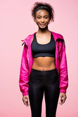attractive african american sportswoman in windbreaker looking at camera and smiling isolated on pink clipart