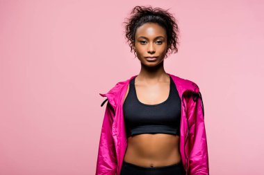 confident attractive african american sportswoman in windbreaker looking at camera isolated on pink with copy space clipart