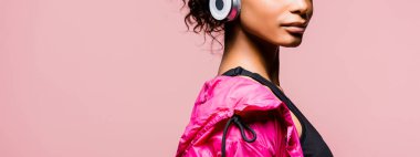 panoramic shot of african american sportswoman in windbreaker and headphones isolated on pink with copy space clipart