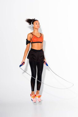athletic african american sportswoman in headphones exercising with skipping rope on white clipart
