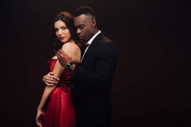 african american man undressing beautiful woman in red dress isolated on black with copy space clipart