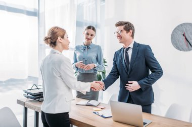 cheerful recruiter shaking hands with woman near colleague in office clipart