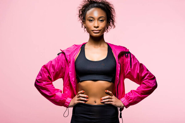 attractive african american sportswoman in windbreaker looking at camera and posing with hands akimbo isolated on pink