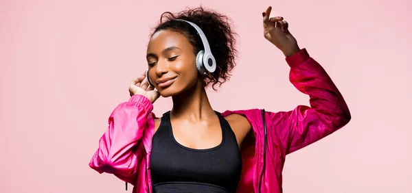 美丽的非洲裔美国女运动员的全景拍摄耳机听音乐和手势孤立在粉红色 — 图库照片