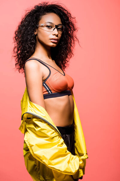 beautiful african american sportswoman in glasses and windbreaker looking at camera isolated on coral