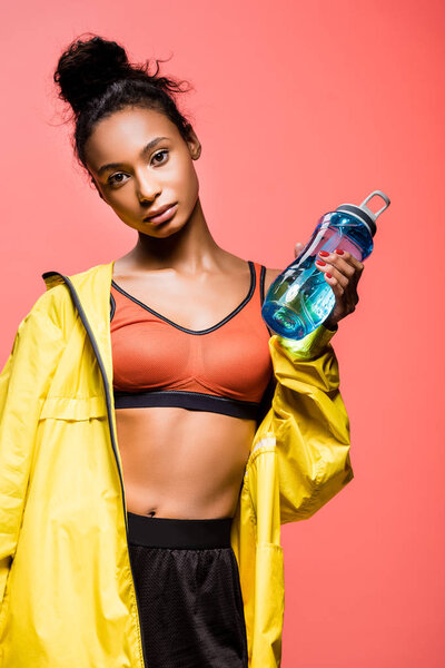 beautiful african american sportswoman with sport bottle looking at camera isolated on coral