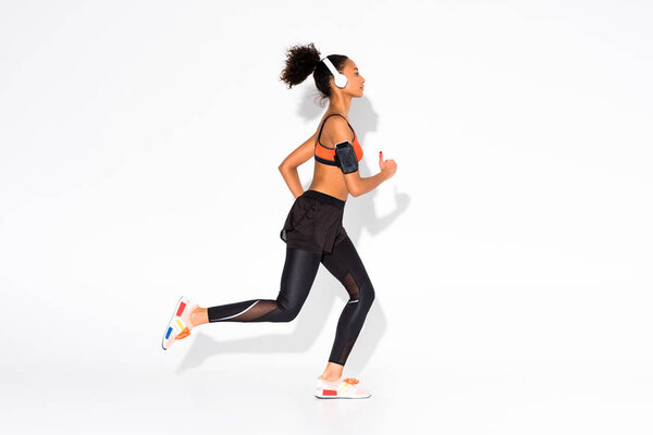athletic african american sportswoman with headphones and smartphone armband running on white