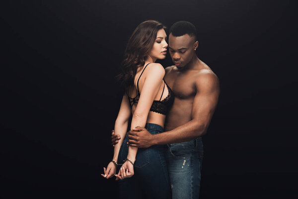 shirtless african american man hugging beautiful sexy woman in handcuffs isolated on black with copy space