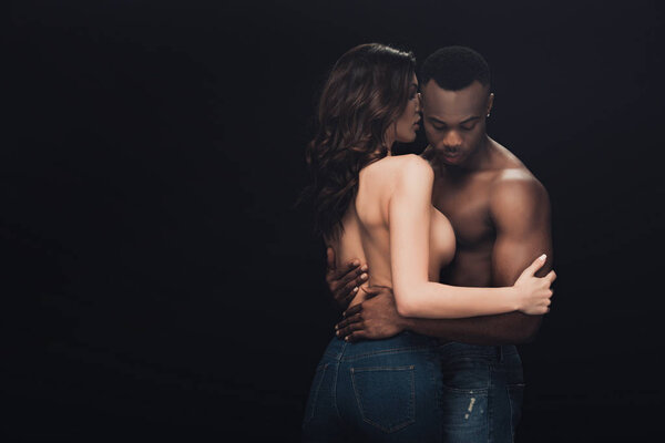 beautiful half-naked interracial couple in denim embracing isolated on black with copy space