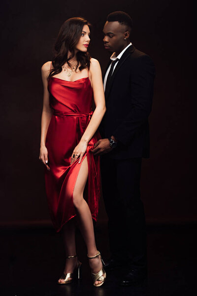 beautiful interracial couple in formal wear looking at each other and posing isolated on black