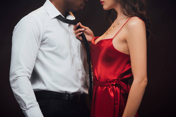 cropped view of woman in red dress holding tie of african american man isolated on black
