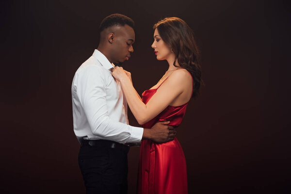 side view of beautiful woman in red dress undressing shirt of african american man isolated on black
