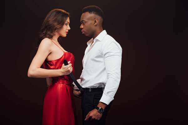 attractive woman in red dress undressing african american man isolated on black with copy space