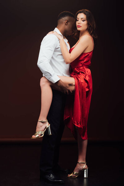 african american man holding leg of beautiful seductive woman in red dress isolated on black