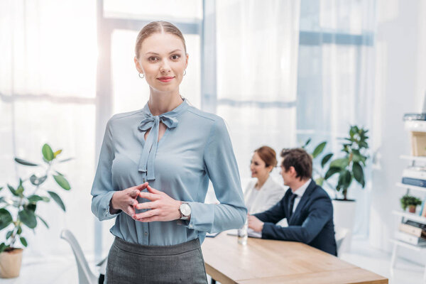cheerful woman standing with clenched hands near coworkers in office 