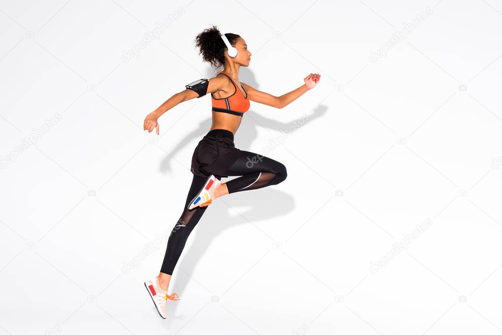 side view of beautiful athletic african american sportswoman in headphones and smartphone armband jumping on white