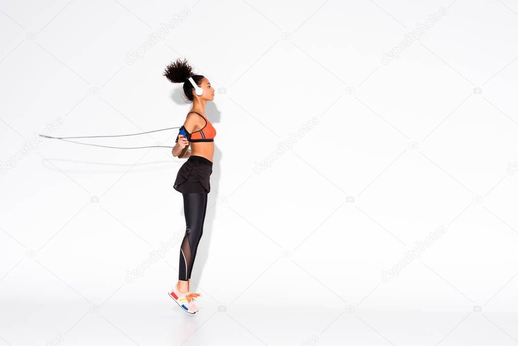 athletic african american sportswoman in headphones exercising with skipping rope on white