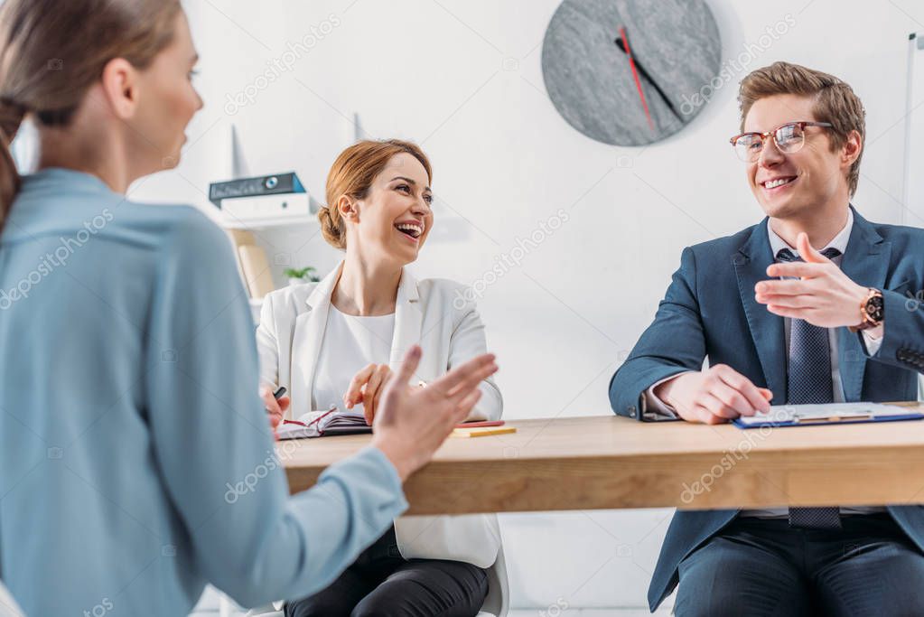 cheerful recruiter looking at coworker in glasses gesturing while speaking with attractive applicant 