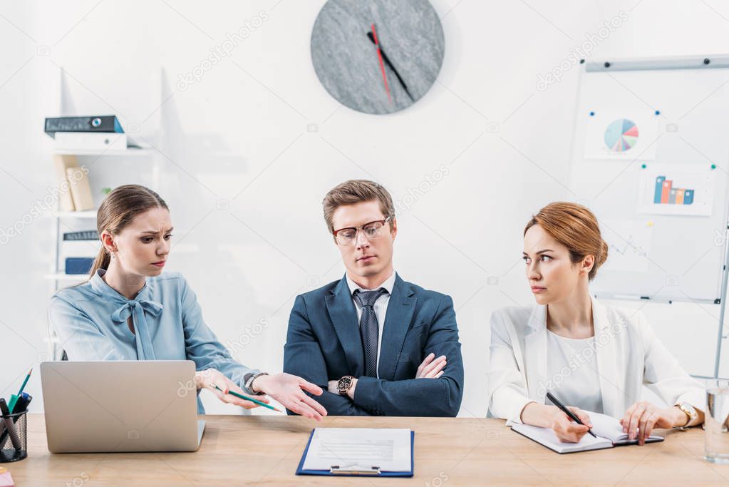 attractive recruiter gesturing near coworker in glasses sitting with crossed arms 