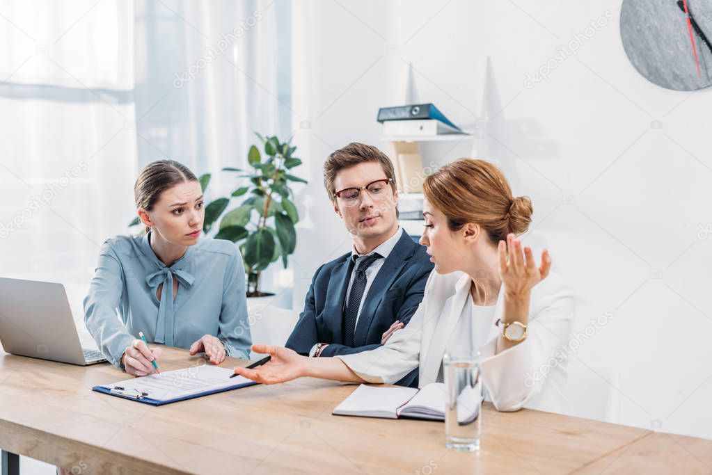 attractive recruiter gesturing near coworker in glasses sitting with crossed arms 
