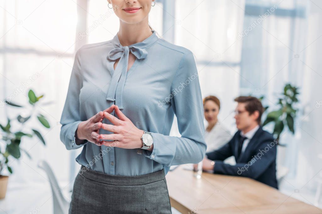 cropped view of cheerful woman standing with clenched hands near coworkers in office 