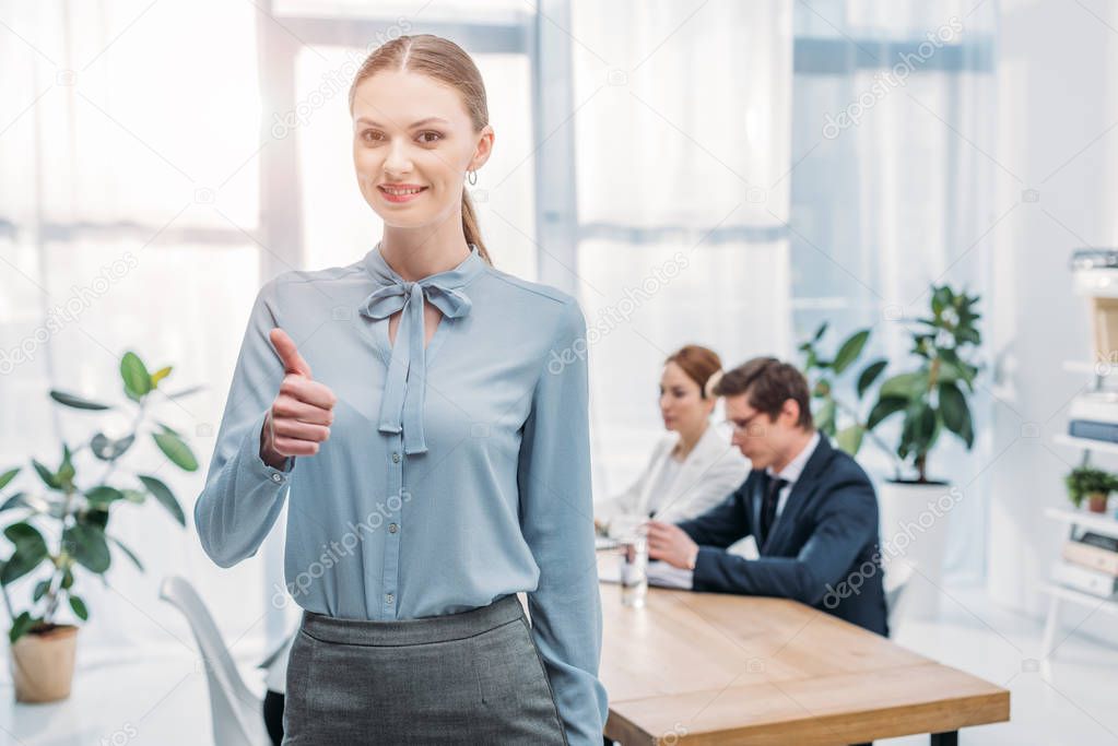 beautiful recruiter showing thumb up while standing near coworkers in office 