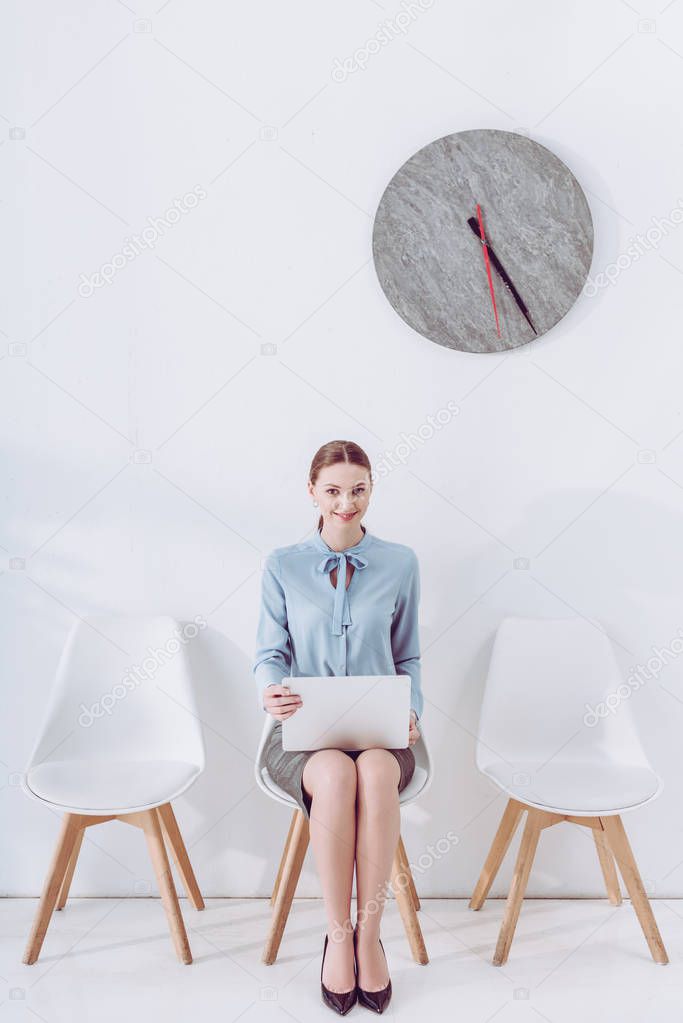 cheerful woman sitting on chair with laptop while waiting job interview 