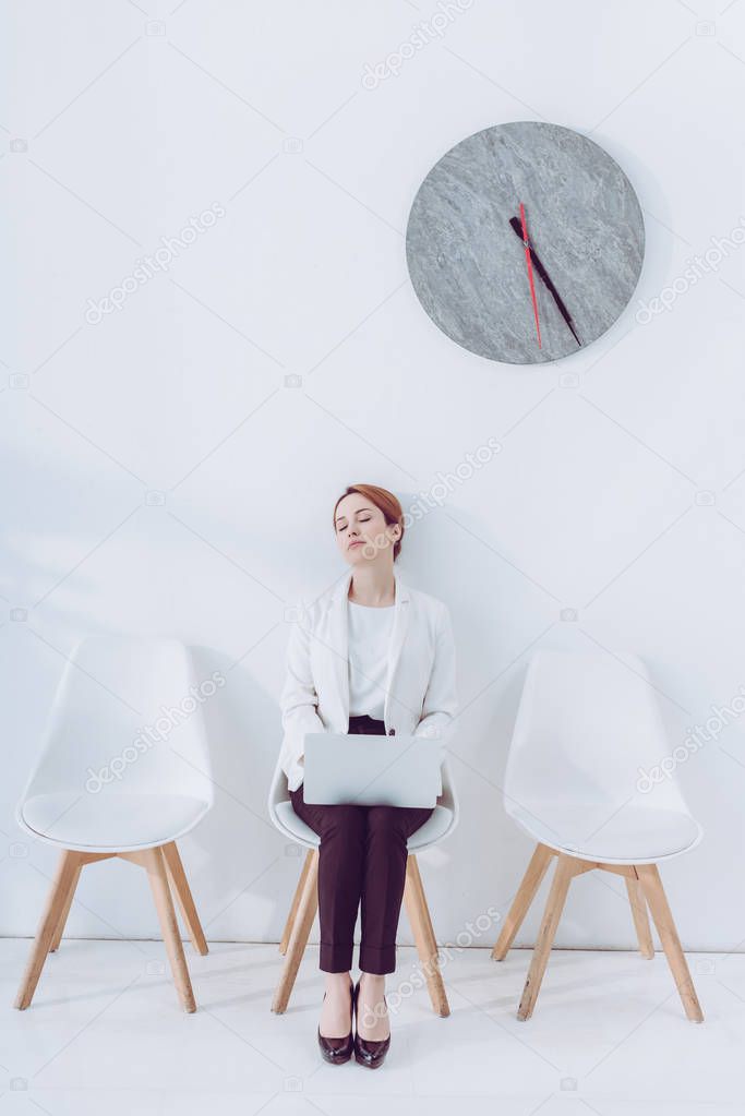 exhausted employee with laptop sleeping on chair near clock 