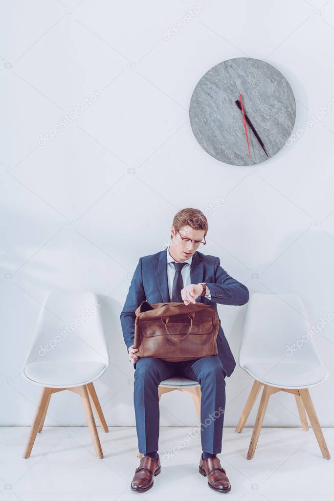 handsome employee in glasses holding briefcase and looking at watch 