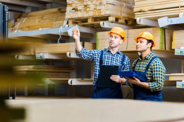 selective focus of two multicultural storehouse workers with clipboards standing in warehouse clipart