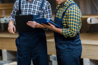 cropped view of two multicultural warehouse workers in uniform holding clipboards clipart