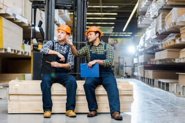 two multicultural workers talking while sitting on plywood in warehouse clipart