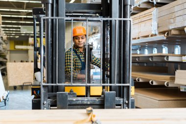 selective focus of handsome, conentrated indian worker sitting in forklift machine in warehouse clipart