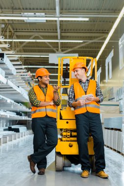 smiling multicultural workers talking while standing near scissor lift  clipart