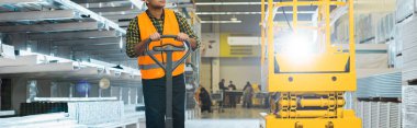 panoramic shot of indian worker in safety vest near pallet jack in warehouse clipart