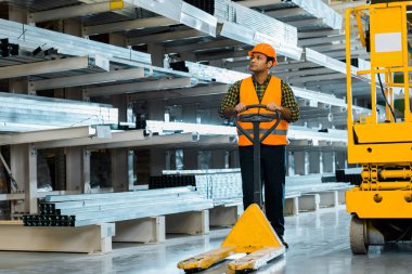 handsome indian warehouse worker standing near pallet jack in warehouse clipart
