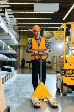 handsome warehouse worker standing with pallet jack near scissor lift clipart