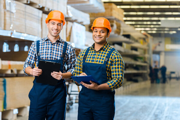 two multicultural workers in uniform smiling and looking at camera in warehouse
