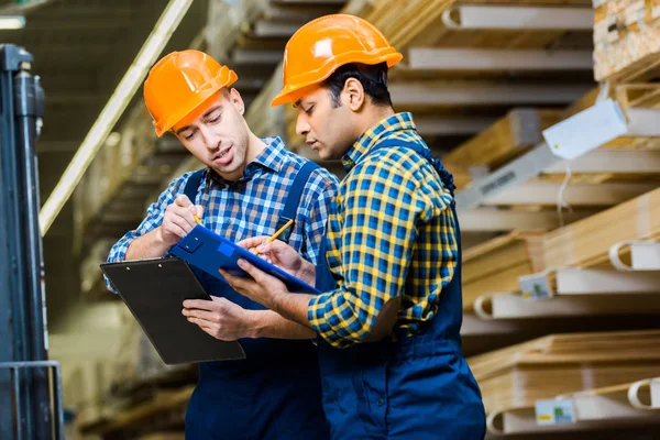 Twee Aandachtige Multiculturele Arbeiders Uniform Helmen Die Klemborden Schrijven — Stockfoto