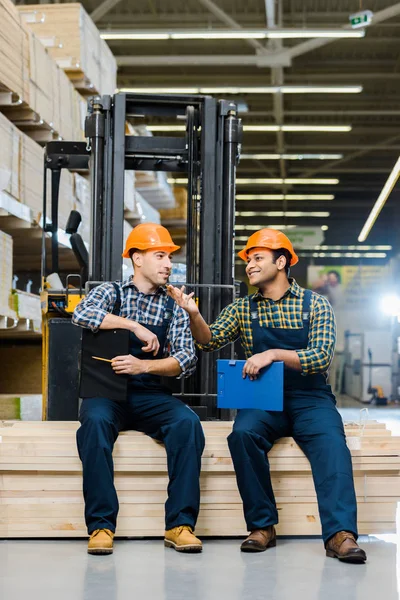 Two Smiling Multicultural Workers Clipboards Talking While Sitting Plywood — Stock Photo, Image
