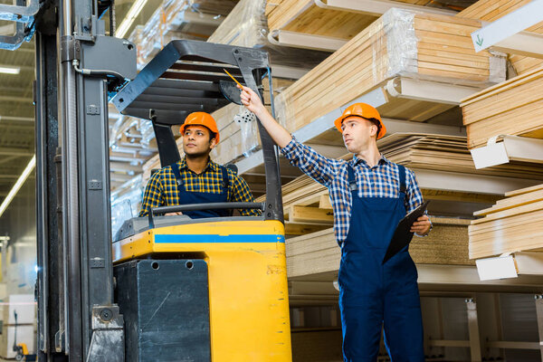 warehouse worker pointing with pencil near inidian colleague sitting in forklift machine