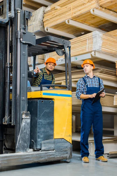 indian worker in forklift machine pointing with finger near colleague with clipboard