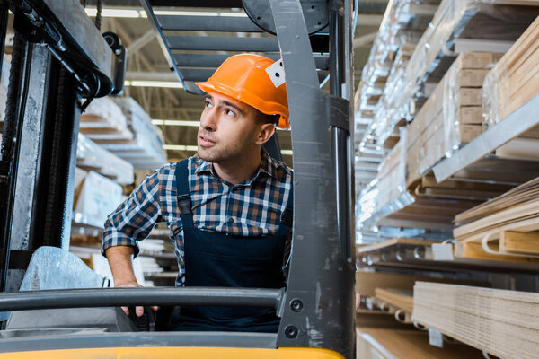 attentive warehouse worker in uniform and helmet operating forklift machine